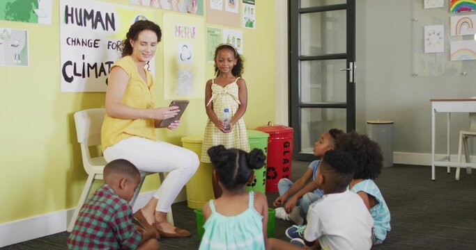 Teacher in yellow blouse guiding six children in semicircle in class with tablet and recycling bins