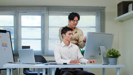 two business people consulting on a project looking at computer monitor screen sitting in the office. Asian male employee Coworker explains colleagues ideas and strategies accountant, financial report