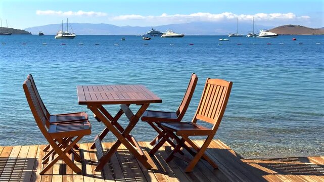 Seaside dining deck with wooden tables and chairs overlooking a beautiful turquoise bay, sailboats, and distant hills under a clear blue sky.