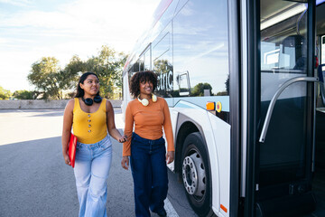 Two young student friends wearing headphones walking towards a public city bus, ready for their daily campus commute