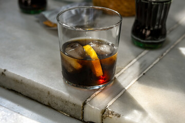 Glass of cola with ice cubes and a slice of lemon on a marble table, illuminated by warm sunlight. The simple composition evokes the relaxed atmosphere of a café terrace in Seville