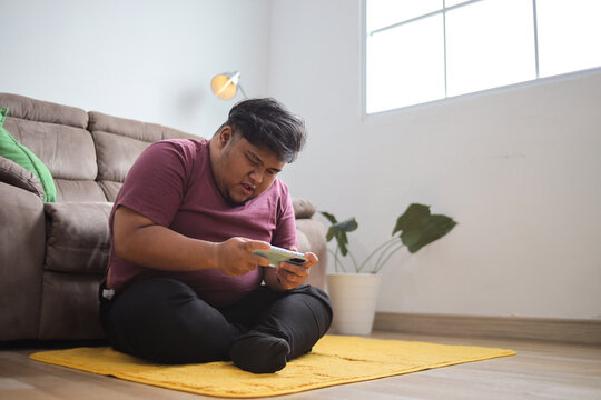 Young Fat Man Enjoying Mobile Game at Home, Sitting on Yellow Carpet in Living Room