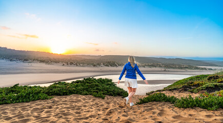 Sightseeing of Algarve coast in Portugal. Middle-aged woman tourist walking on path overlooking beautiful sandy beach and river Amoreira during sunrise in summer. Back view	