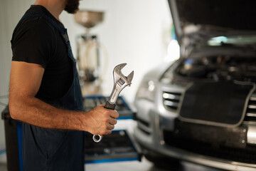 A car mechanic standing in a repair shop uses a wrench to fix a vehicle. The shop is well-lit with tools and equipment in the background.