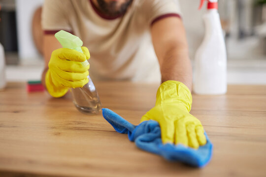 A person wearing yellow gloves sprays a cleaner on a wooden kitchen surface and wipes it with a blue cloth. The scene shows a clean and organized home environment.