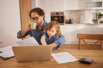 A father is balancing work and family as he enjoys a cup of coffee with his young daughter beside him, engaging with documents at a kitchen table during the afternoon.