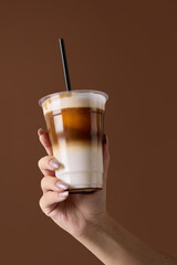 Hand holding iced latte in plastic cup with straw on brown background. Concept of barista craft, beverage composition, product presentation for cafe advertising and packaging design.