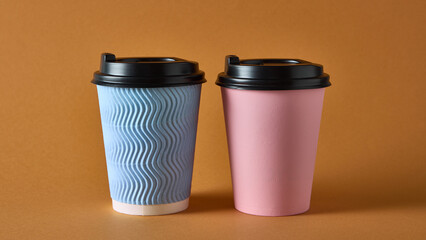 Blue textured and pink takeaway coffee cups with black lids on orange background. Concept of cafe product mockup, packaging design, branding for beverage and catering industry visuals.