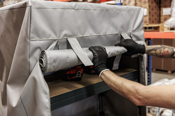 A person with gloves rolls up a large gray industrial fabric cover, securing it with Velcro straps...