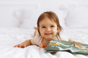 Smiling baby lying on bed and playing with colorful soft book. Joyful moment expressing learning, curiosity, and happy childhood at home in bright light