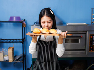 Asian beautiful woman bakers wearing striped aprons holding showing fresh baked salted butter roll bread on wooden tray smells good in bakery kitchen making homemade pastry baking handcrafted concept.
