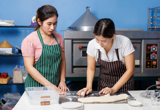 Two asian woman baker wearing striped apron preparing round fresh dough balls on metal plate table for bread pastry making, Homemade bakery process showing soft dough texture, step baking in kitchen.