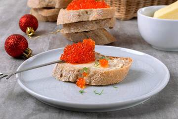 Red caviar and butter on a slice of baguette on a gray table. Basket with rye bread in the background. Festive snack
