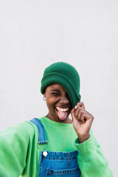 Cheerful woman wearing green knit hat sticking out tongue in front of white wall
