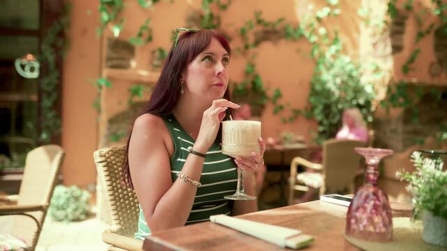 Gorgeous young lady relaxing at an al fresco cafe table, savoring a cool iced coffee frappe under the sun in the bustling city