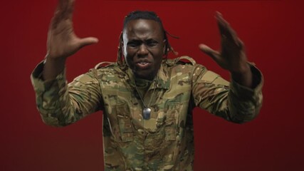 Man, young in camouflage with dogtags gesturing with hands in red studio; frustration.