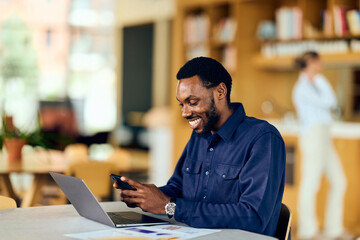 Fototapeta premium African American Man Working On Laptop And Phone In Coffee Shop, Smiling And Focused, Productive