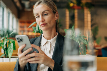 A middle-aged blonde woman in a restaurant checks her smartphone while waiting for her lunch order.