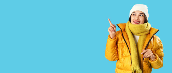 Young Asian woman in winter clothes pointing at something on blue background
