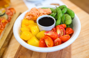 Colorful poke bowl with fresh salmon, mango cubes, edamame and cherry tomatoes arranged around soy sauce for a vibrant and balanced meal ready to eat. Healthy and natural fast food concept.