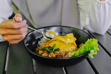 Close-up shot of hands using a spoon and fork to eat a bowl of savory rice dish topped with soft chiffon-style egg and meat