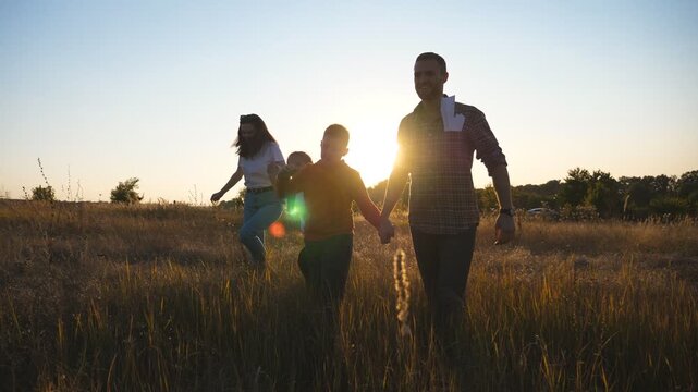 Mom and dad with two children going among summer meadow and enjoy to spending time together. Young parents with small kids walk through grass field holding hands of each other at sunset. Slow motion