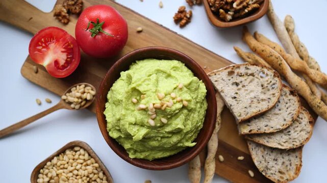 Top view of a delicious and healthy appetizer featuring a bowl of fresh avocado guacamole dip sprinkled with pine nuts, served with tomatoes, walnuts, and whole grain breadsticks