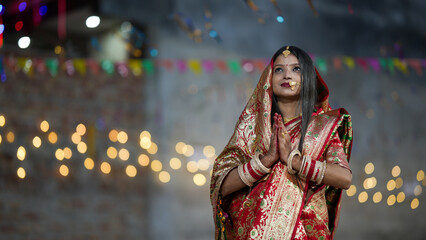 Newly married Indian woman in red saree doing namaskar with diya lights and bokeh festival background, symbolizing devotion and grace.