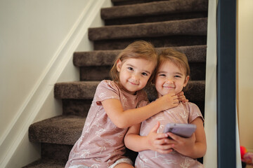 Two preschool age twin sisters hugging and using smartphone on stairs at home