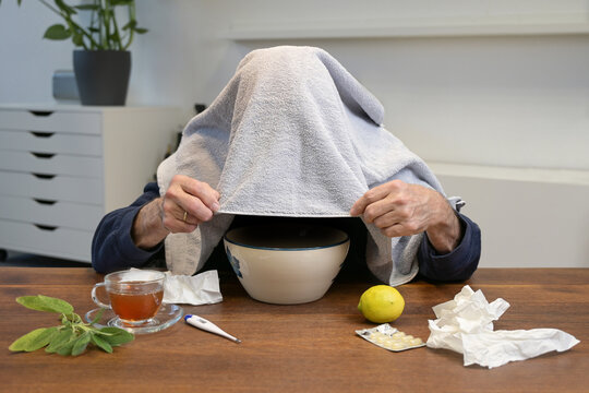 Man doing steam inhalation over a bowl with a towel over his head at a table with many home remedy treatments, lemon, tea, fever thermometer and used tissues, cold and flu health concept