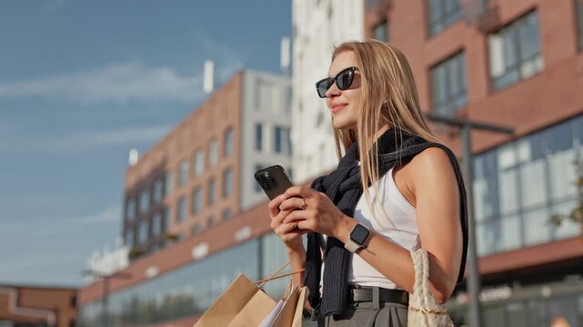 Woman with long hair wearing sunglasses and standing near buildings. Holding smartphone in both hands and looking at screen. Checking messages or browsing social media while carrying shopping bags.