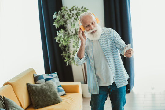 Happy senior man with white beard listens to music on bright orange headphones while holding a smartphone at home smiling