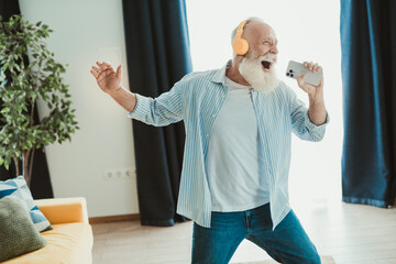 Senior man dancing at home wearing headphones with phone in hand expresses joy and energy in a bright living room