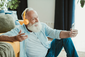 Senior man at home enjoys music with headphones taking a selfie on the couch in a cozy living room for a relaxed joyful moment