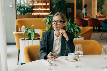 Blond middle-aged woman in a blazer sits in a stylish restaurant, thinking while using her phone