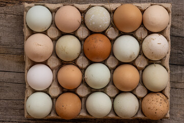 Closeup flat lay of carton full of organic eggs on old wood desk. Natural product differ of colors and surface pattern with dirt on shell and small pieces of hay among emphasized rural simplicity.