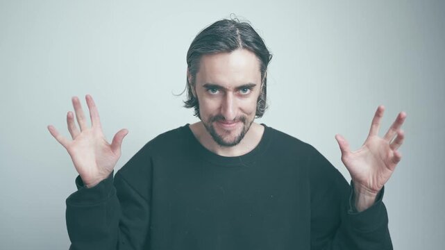 Cunning Smile and Cringy Dance of Man with Medium-Length Wavy Hair and Beard in Black Sweatshirt Against Soft Pale Background
