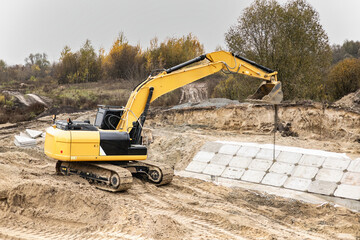 Excavator digs and moves heavy materials on a construction site surrounded by trees under cloudy skies. The ground is sandy and marked with tracks