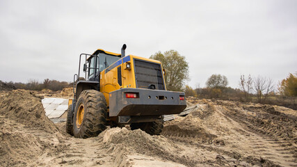 A large yellow tractor is digging in sandy soil at a construction site. The cloudy sky and autumn trees create a somber backdrop for the activity