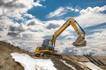 Excavator is digging into a mound of dirt at a construction site. The sky is filled with clouds, and equipment is visible in the background