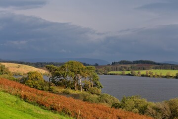 A scenic landscape features a tranquil lake surrounded by rolling hills under a cloudy sky in Killington Lake - Kendal - Lake District - UK