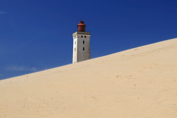 Landscape photo with a view of the Rubjerg Knude lighthouse (also known as the "Lighthouse in the Dunes") with sand dunes in the foreground against a blue sky with clouds near Lokken in Denmark