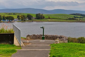 A scenic overlook includes a telescope at a lake, surrounded by green grass. Mountains form the background in Killington Lake - Kendal - Lake District - UK