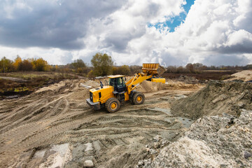 A yellow loader operates on a construction site, moving dirt and gravel while clouds gather overhead in a remote area during daytime