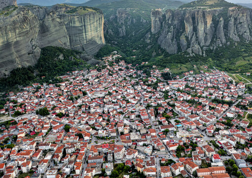 Aerial view of clustered red-roofed houses nestled beneath the imposing cliffs of Meteora, a symphony of human construction against a backdrop of natural majesty, Kalabaka, Trikala, Greece.