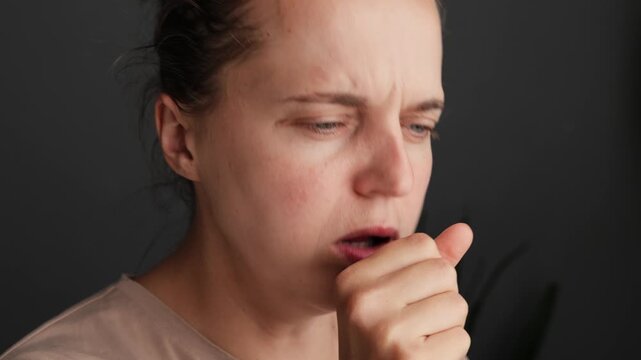 A woman sits indoors, experiencing discomfort as she coughs softly into her hand. The room is dimly lit, and her expression shows concern
