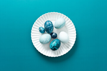 Colorful blue Easter eggs are artistically arranged on a white plate resting on a vibrant teal...
