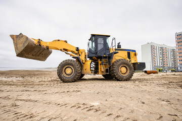 A large loader is working on a sandy area next to residential buildings. The overcast sky adds a muted backdrop to the scene
