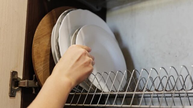 Someone is organizing clean dishes in a kitchen cabinet, carefully placing one on a shelf while surrounded by other dishware in the evening light