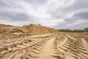Deep tracks from heavy machinery cut through the sandy ground on a cloudy day. A large pile of sand rises in the background, hinting at ongoing construction